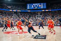 Jogo de basquetebol num pavilhão com jogadores em acção e público nas bancadas